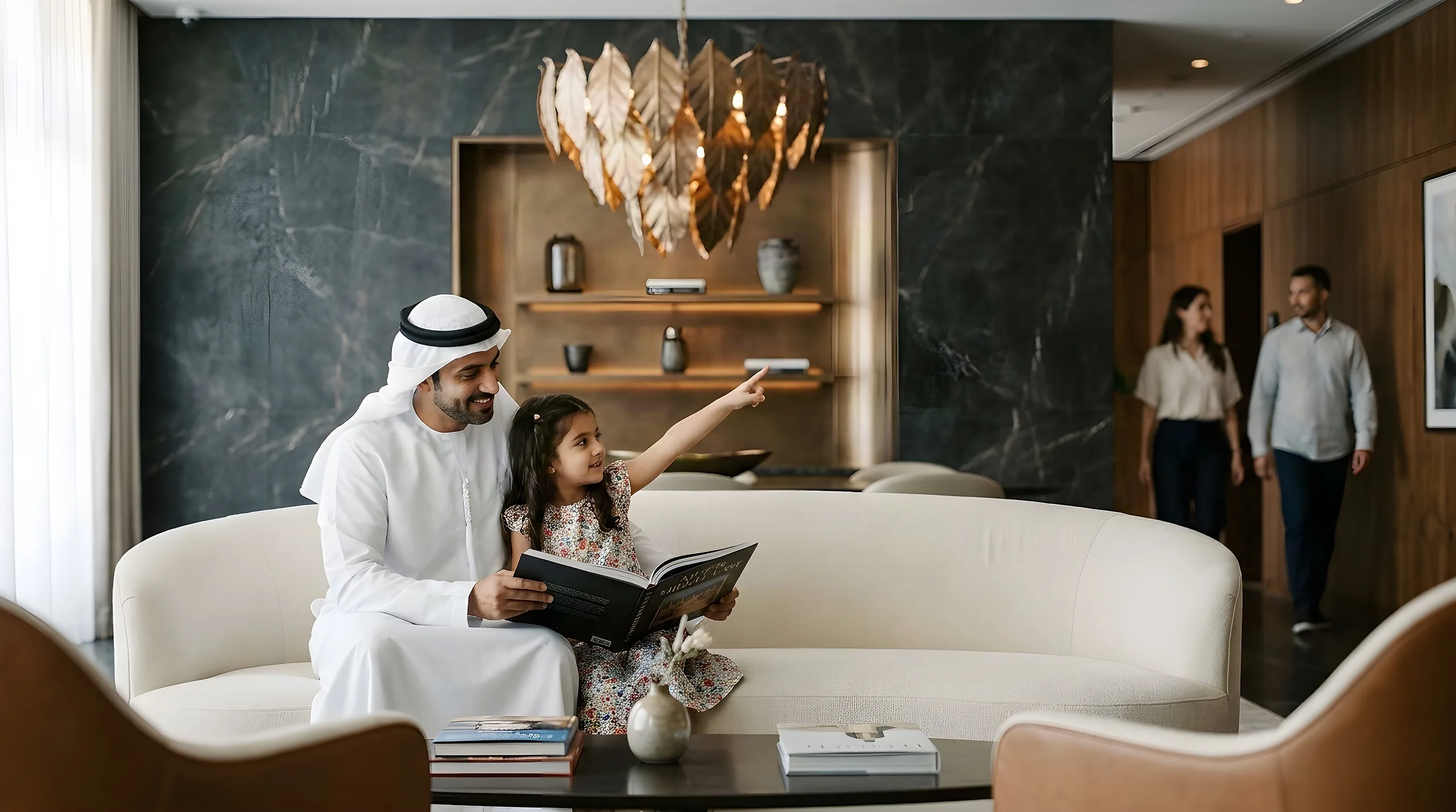 A father in a white kandura reads a picture book with his daughter on a curved cream sofa under a bronze leaf chandelier, a couple walking in behind them