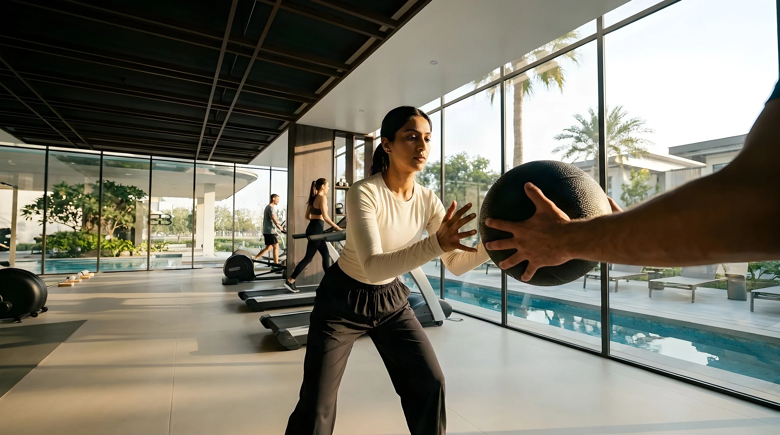 A woman catches a medicine ball by the gym’s glass wall, treadmills behind her and the pool outside in low sun